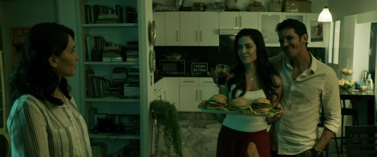 Human Animals (2020) movie still: medium — A residential kitchen features white cabinetry, shelves filled with books, and a green tint. One woman…