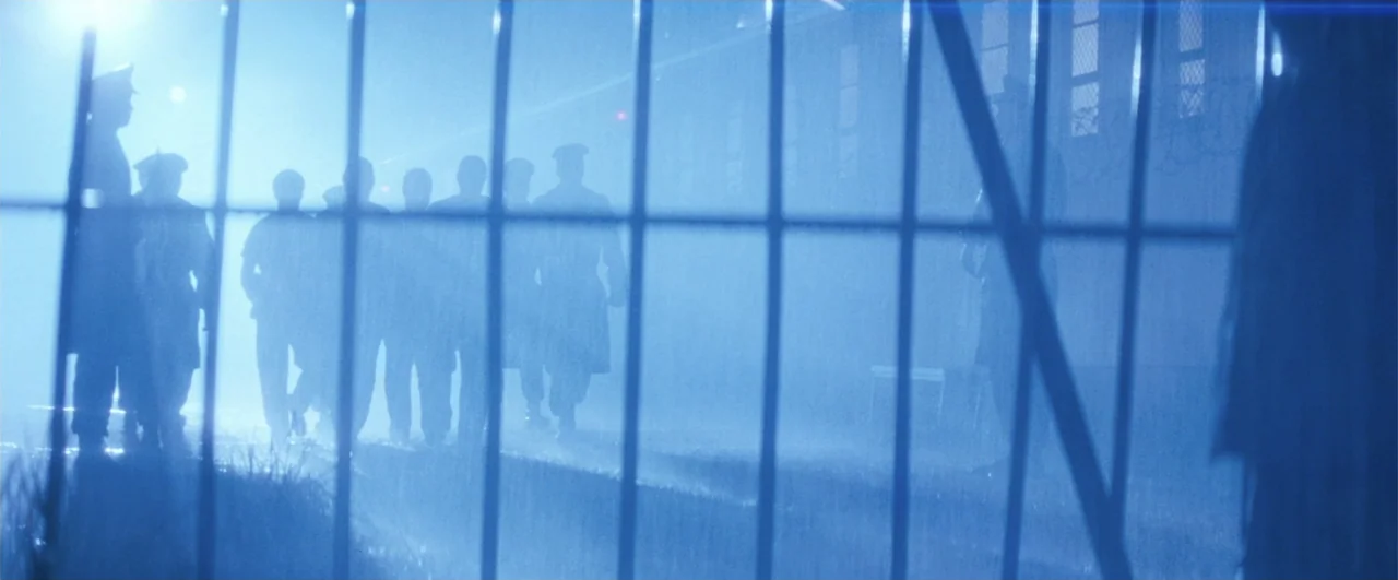 Lethal Weapon 4 (1998) movie still: wide — A group of uniformed men walks behind a tall chain-link fence at night. Thick blue-tinted fog obscures…