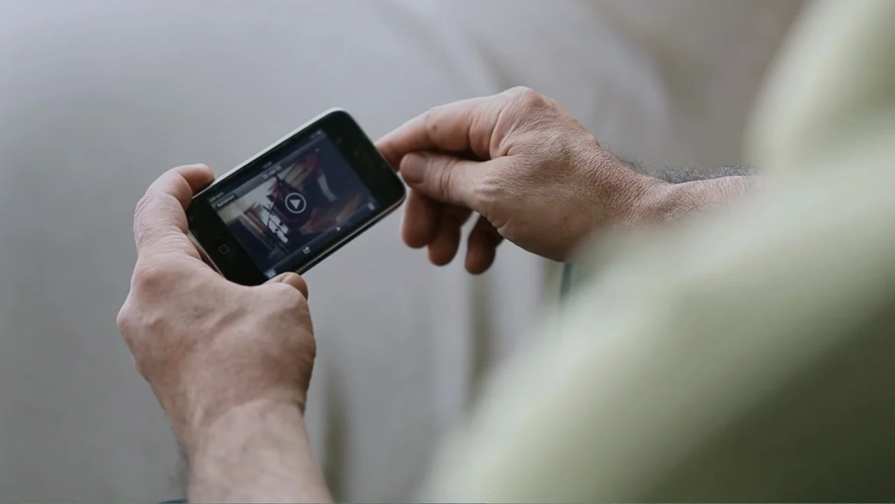 Closed Curtain (2013) movie still: closeup — A close-up view shows a person holding a smartphone indoors. The person interacts with the mobile…