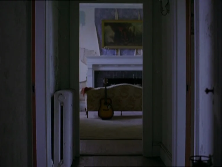 Last Days (2005) movie still: wide — A dimly lit narrow corridor leads into a brighter room with peeling wallpaper. An acoustic guitar stands…