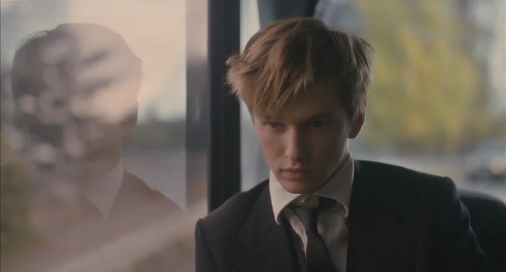 Restless (2011) movie still: medium — A person sits inside a vehicle next to a window showing a blurred exterior. The young man gazes ahead while…