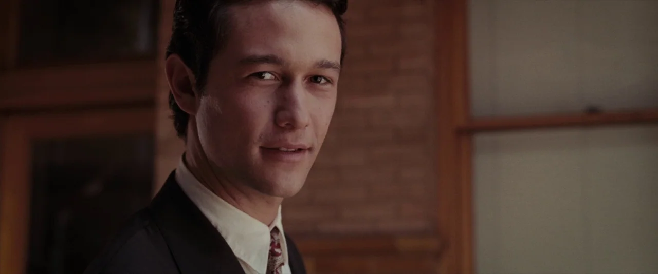500 Days of Summer (2009) movie still: closeup — A brick building facade stands behind a man in a dark suit. The man looks toward the camera while…