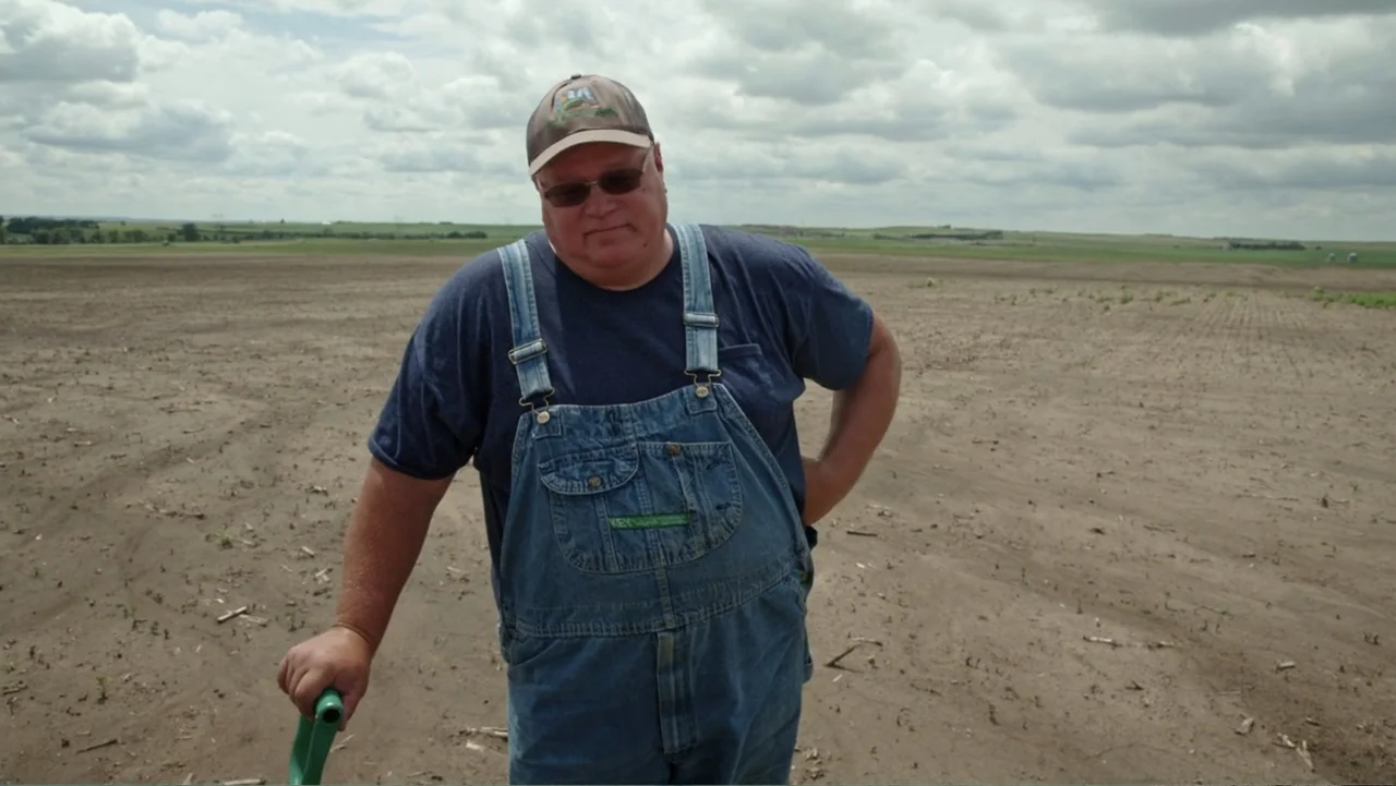 Common Ground (2000) movie still: medium — A vast, flat agricultural field under a sky filled with scattered white clouds. A middle-aged man stands…