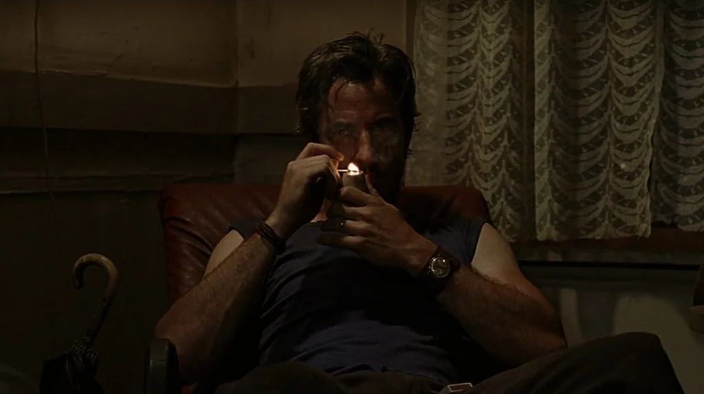 Undertow (2004) movie still: medium — A dimly lit room with lace curtains visible behind a man sitting in a chair. The man lights a pipe while…
