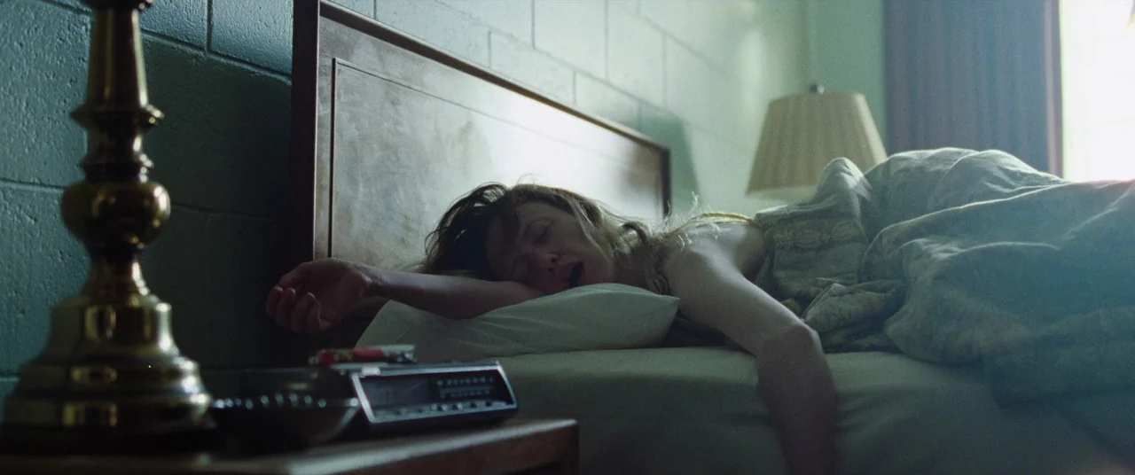 To Leslie (2022) movie still: medium — A modest motel room features concrete block walls and a simple wood headboard. A woman with disheveled hair…