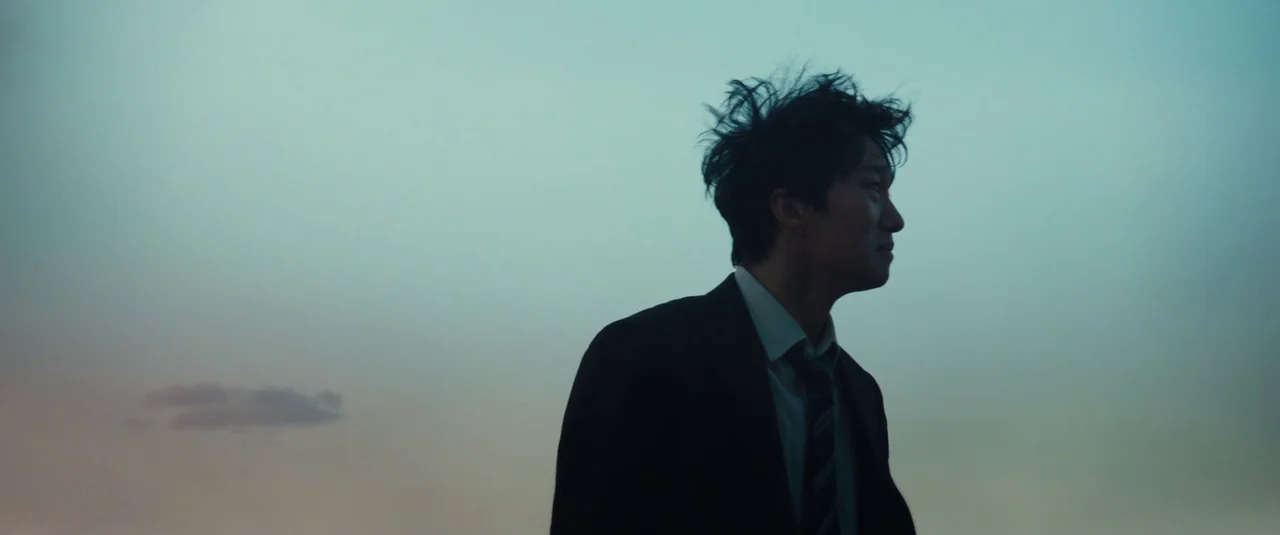 Decision To Leave (2022) movie still: medium — A man in a dark suit stands alone against a vast, overcast twilight sky. He appears in silhouette as…
