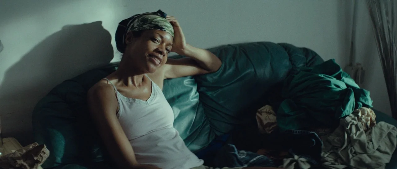 Moonlight (2016) movie still: medium — A woman sits on a teal sofa cluttered with pile of folded laundry. She rests her arm on the back of the sofa…