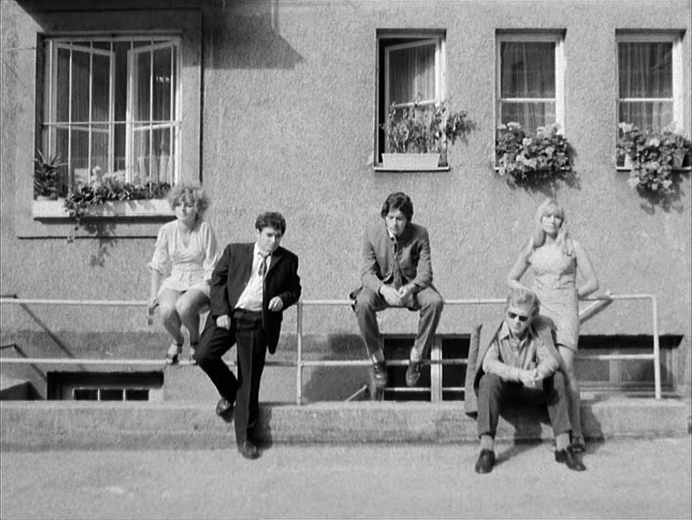 Katzelmacher (1969) movie still: fullbody — A group of five people poses in front of a textured gray building facade. They sit or stand leaning…