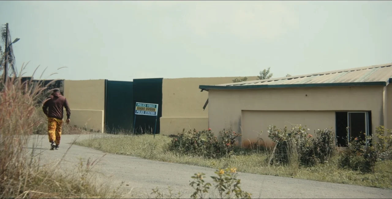 Project Assault (2025) movie still: wide — A paved path leads to a walled compound featuring a sign for a police station. A solitary man in a hooded…