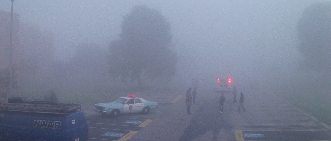 Halloween II (1981) movie still: wide — A paved lot is shrouded in heavy, opaque fog during the night. Several figures gather near a parked police…
