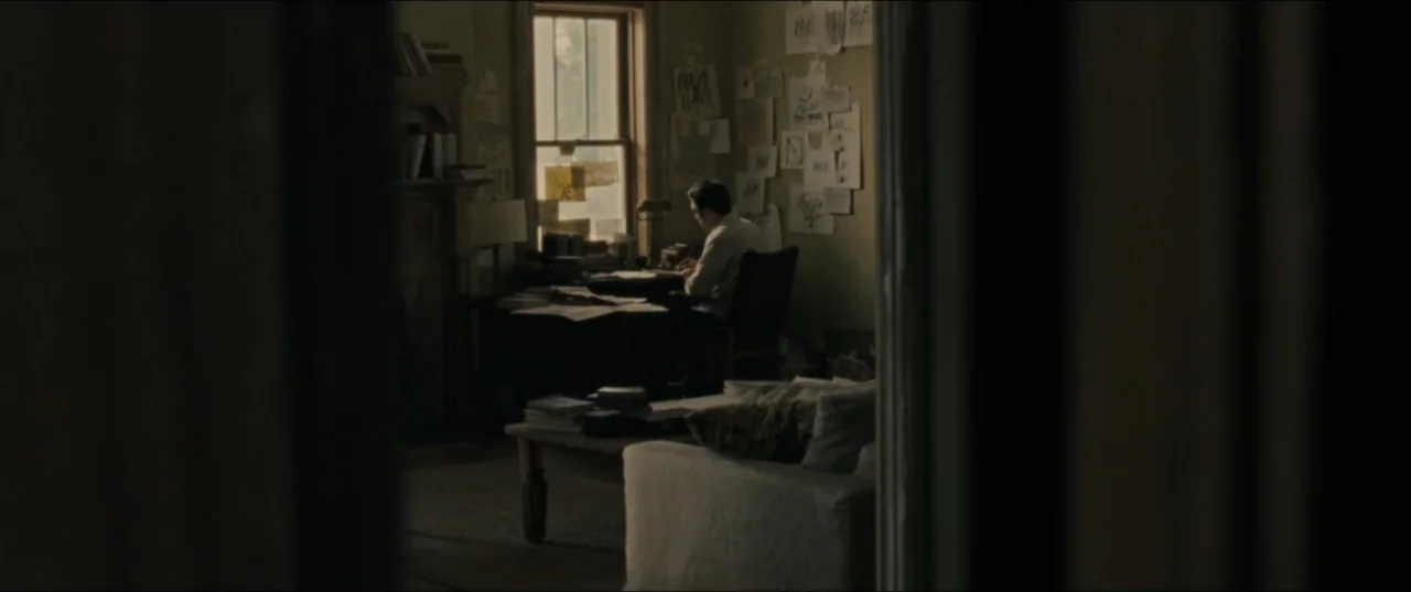 mother! (2017) movie still: wide — A dimly lit room contains a desk, a sofa, and walls covered in scattered notes. A man sits at the desk by the…