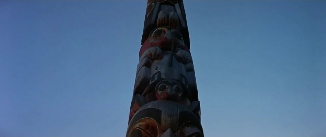 The Parallax View (1974) movie still: closeup — A vertical totem pole with animal carvings stands against a clear twilight sky. The wooden sculpture…