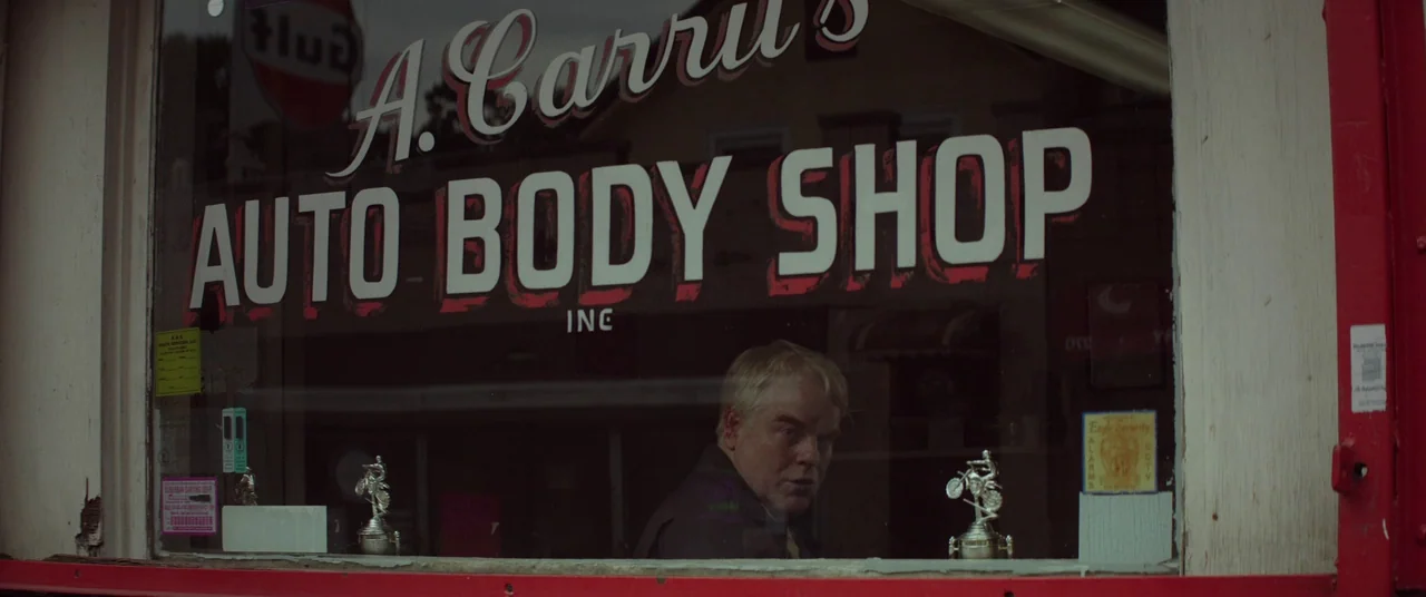 God’s Pocket (2014) movie still: medium — A glass storefront displays the painted text for an auto body shop. A man peers out from behind the…