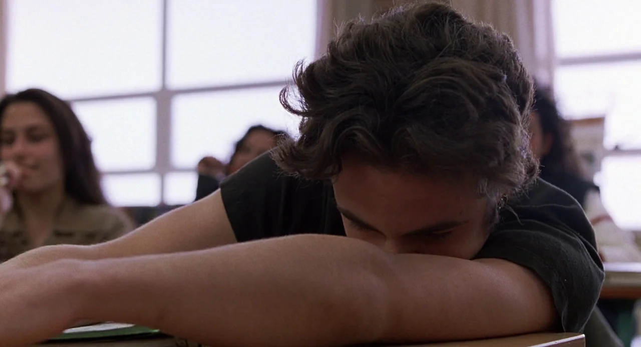 To Die For (1995) movie still: closeup — A high school classroom with large windows in the back and multiple students present. A teenager rests his…