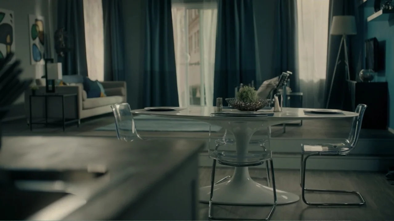 Sightless (2017) movie still: wide — A modern dining room features a white tulip table flanked by transparent plastic chairs. The space is…