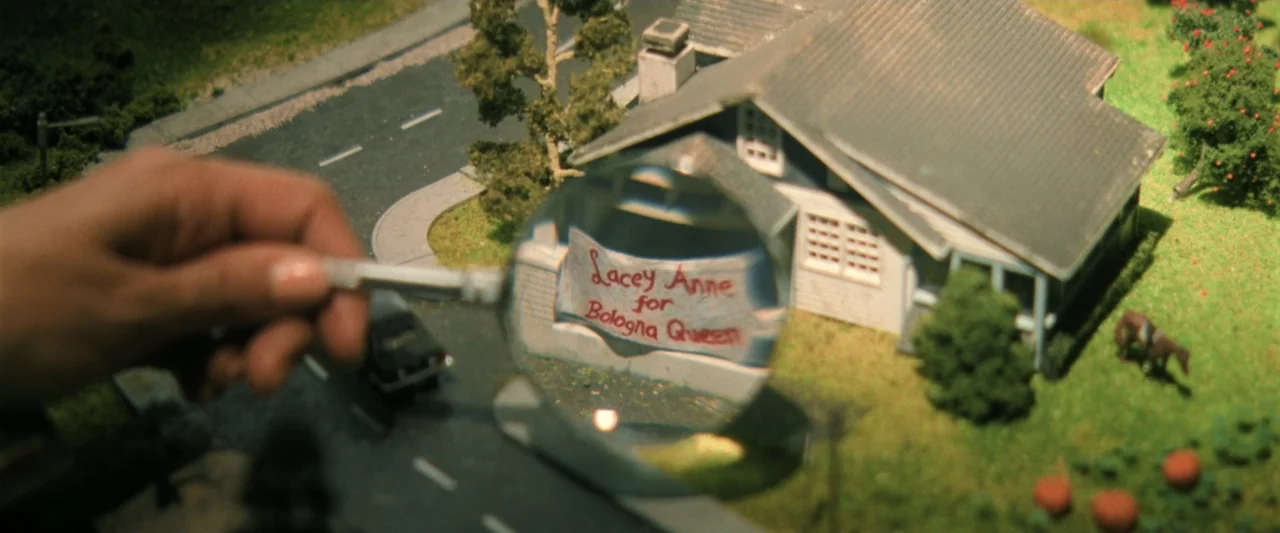 Holland (2025) movie still: medium — A close-up view shows a hand holding a magnifying glass over a suburban diorama. Through the lens, the text on…