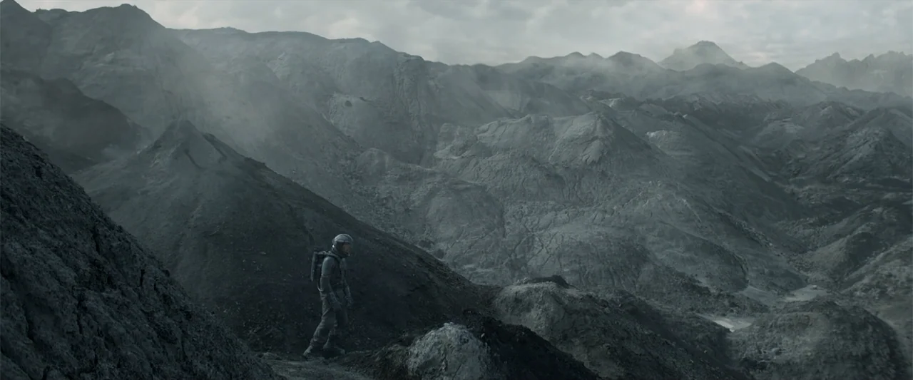 DISTANT (2026) movie still: wide — A vast, desolate mountain range with grey volcanic rock and a cloudy overcast sky. An astronaut in a full…