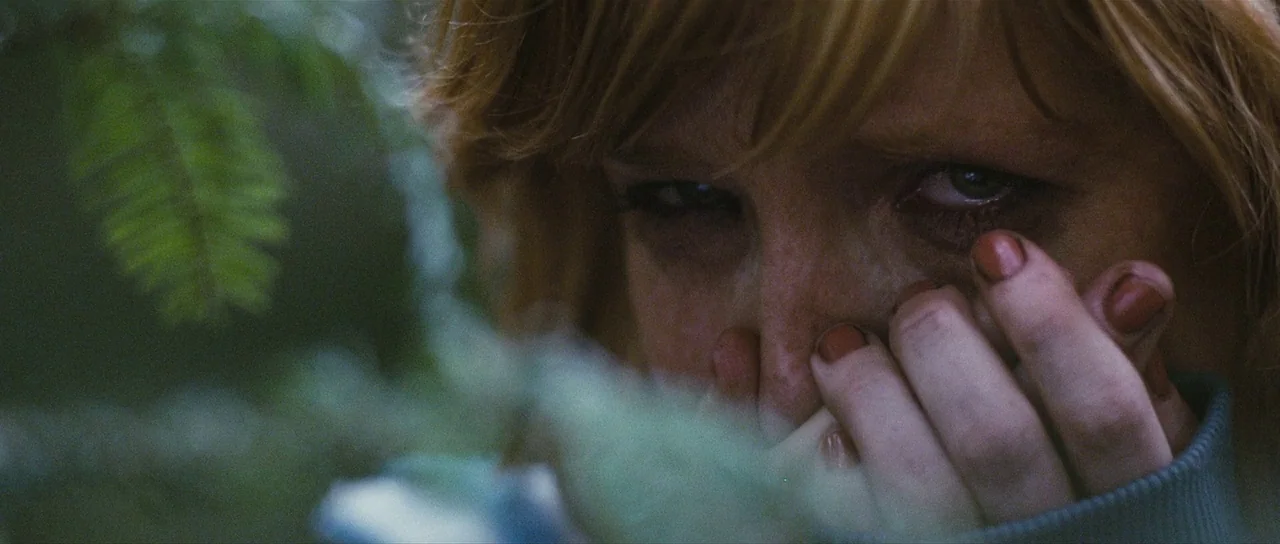 Eden Lake (2008) movie still: closeup — A dense forest environment with evergreen branches in the foreground obscuring the view. A woman with red…