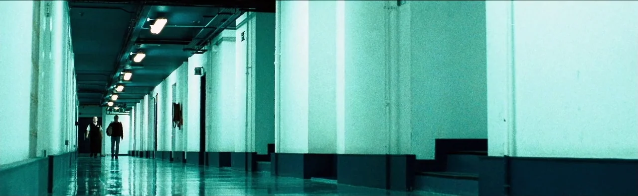 London Boulevard (2010) movie still: wide — A long, symmetrical hospital hallway features white walls with dark wainscoting and polished floors. Two…