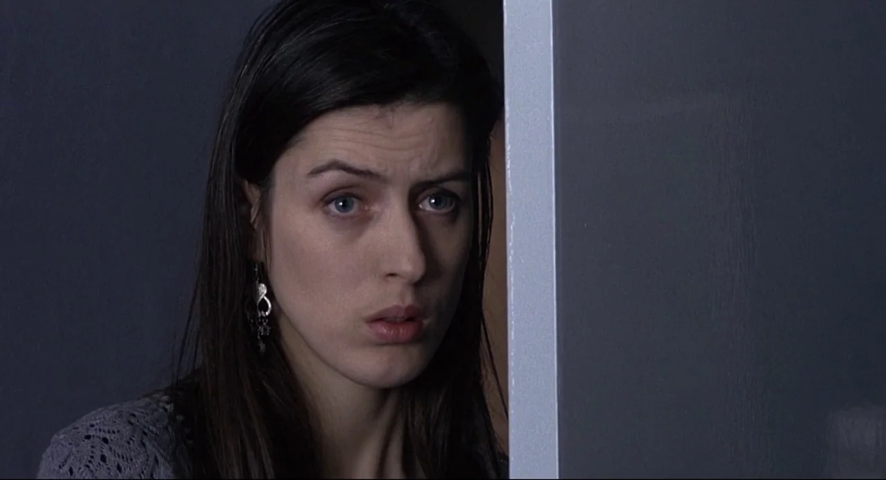 Naked (1993) movie still: closeup — A woman stands behind a doorframe looking into an adjacent room. The lighting is low-contrast and cool-toned.