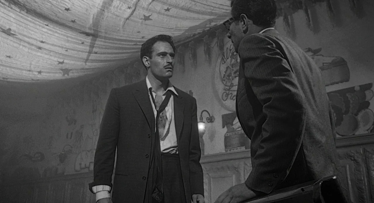 Touch of Evil (1958) movie still: medium — A dimly lit tavern with a star-patterned fabric draped across the ceiling. Two men in formal suits are…