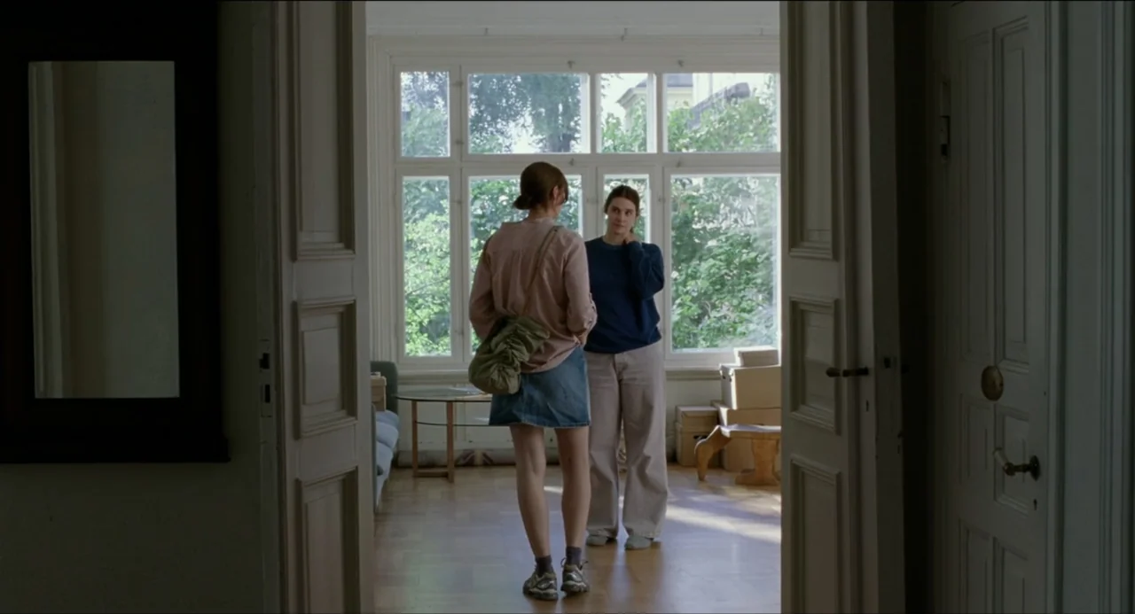 Sentimental Value (2025) movie still: fullbody — A sunlit residential interior with large white-framed windows overlooks trees outside. Two women…