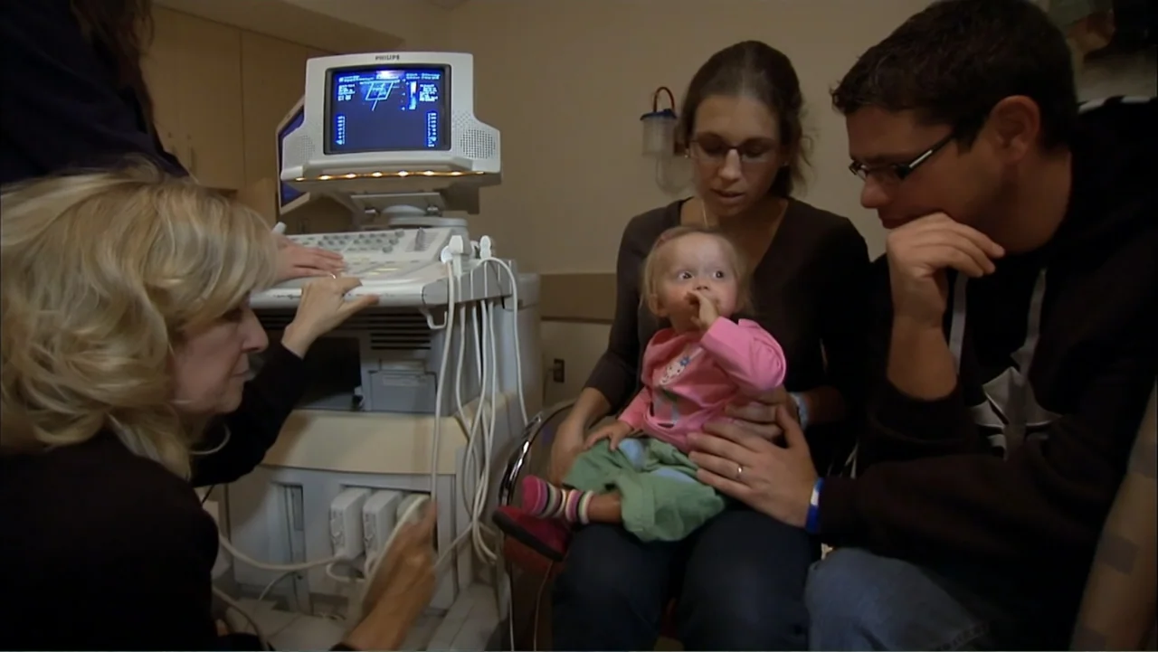 Life According to Sam (2013) movie still: medium — A medical office contains a large ultrasound machine with a glowing screen display. A mother…