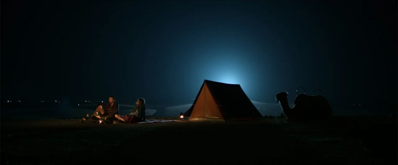An Endless Wedding (2025) movie still: wide — A vast desert expanse at night features a small tent and a resting camel. A man and woman sit together…