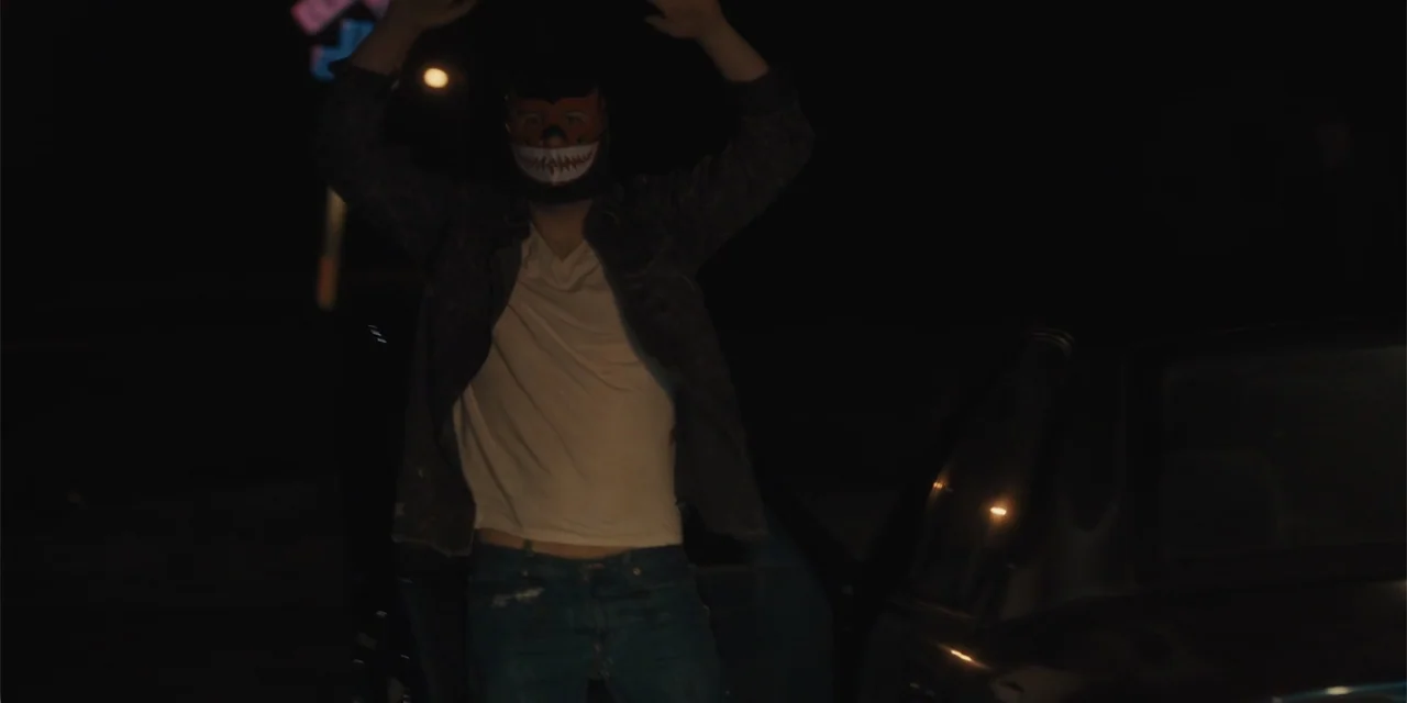 All Roads to Pearla (2019) movie still: medium — A dark outdoor setting at night with the side of a vehicle visible. A man wearing a skull mask…