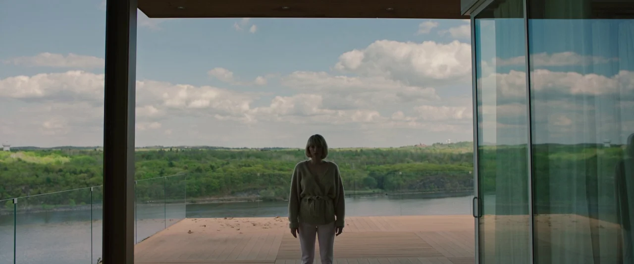 Swallow (2020) movie still: wide — A woman stands on a wooden balcony overlooking a wide river and dense forest. The balcony is bordered by a clear…