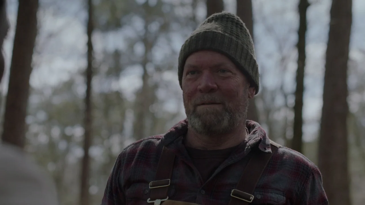 Die Hart (2023) movie still: medium — A man stands within a densely packed forest filled with tall, mature trees. He wears outdoor work clothing…