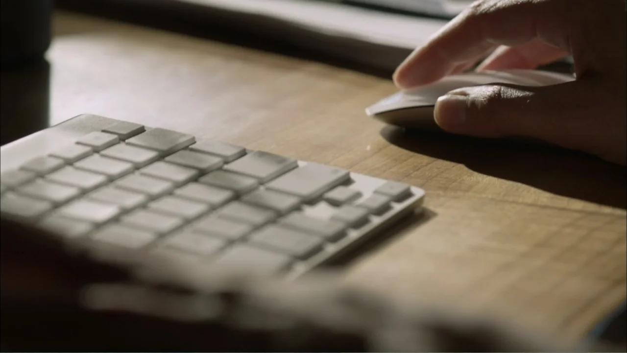 Dark Skies (2013) movie still: closeup — A wooden desktop features a white keyboard and a computer mouse in soft light. A human hand rests on the…