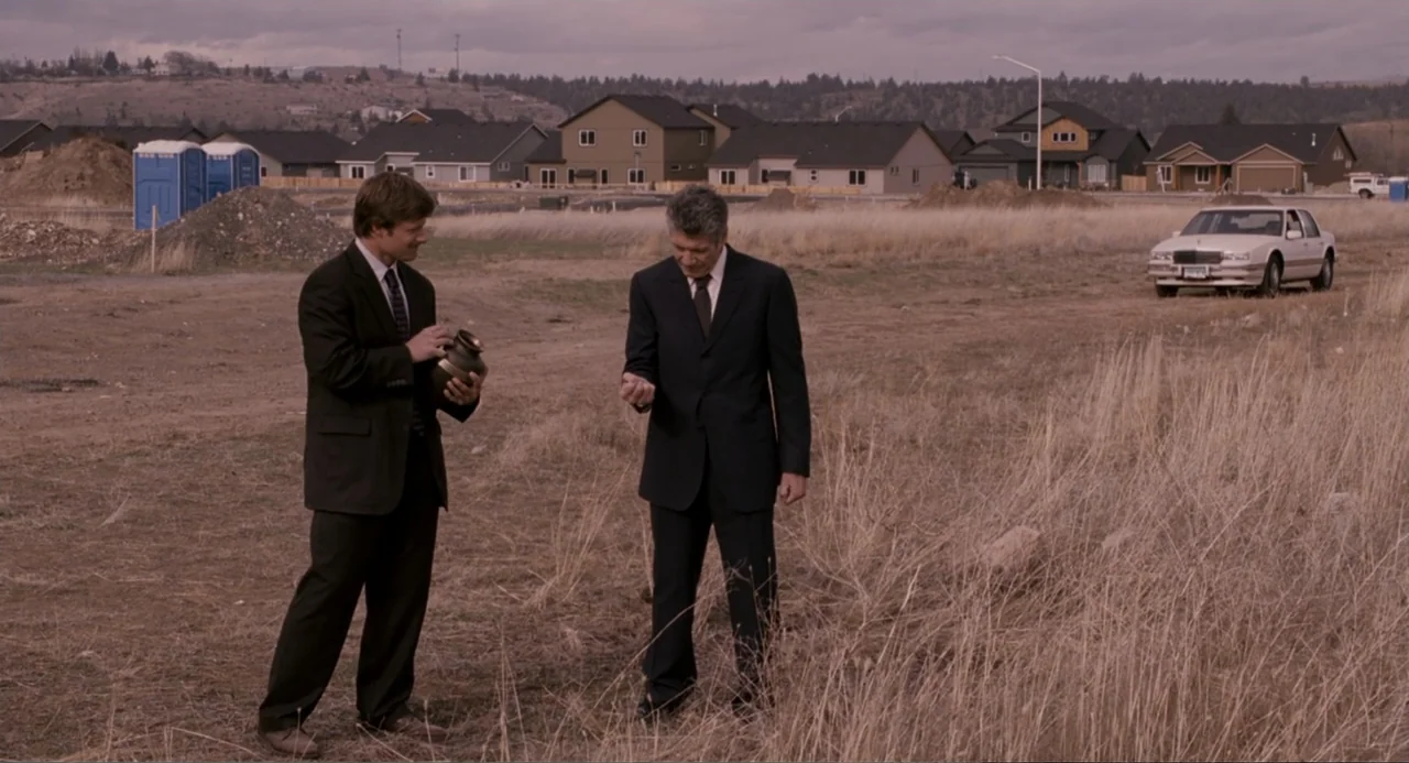 Management (2009) movie still: fullbody — An expanse of dry, brown grass borders a suburban housing development under overcast skies. Two men in…