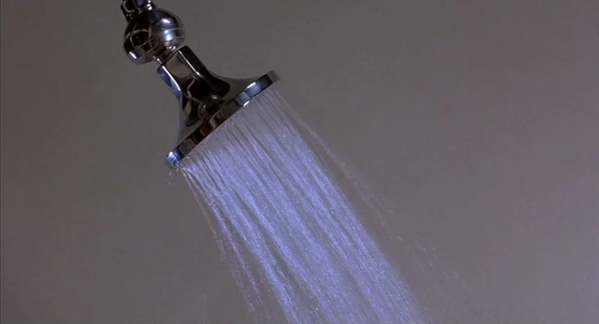 Psycho 2 (1983) movie still: closeup — A metallic showerhead is mounted on a plain, light grey wall. A steady stream of water sprays downward from…