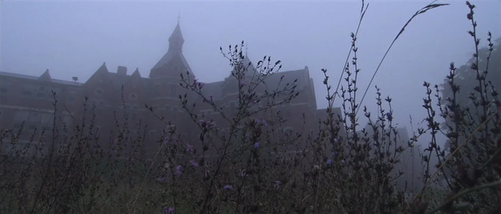 Session 9 (2001) movie still: establishing — An old, multi-story brick institutional building is shrouded in heavy morning fog. Dense wildflowers…