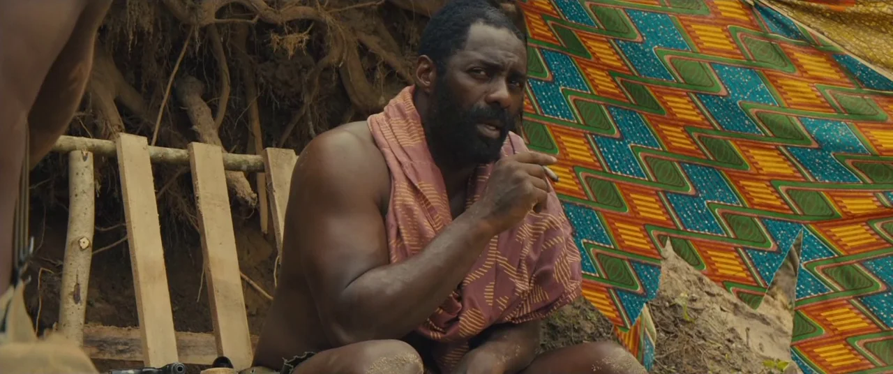 Beasts of No Nation (2015) movie still: medium — A man sits outdoors against a backdrop of exposed earth and patterned cloth fabric. He holds a…