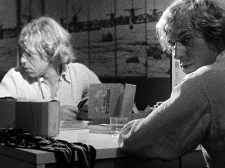 Alice in the Cities (1974) movie still: medium — An interior space features two men sitting at a desk with stacked photo boxes. A large screen…