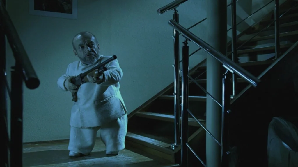 The Ugly Swans (2006) movie still: fullbody — An interior staircase with wooden steps and a metallic silver handrail sits at night. A short man…