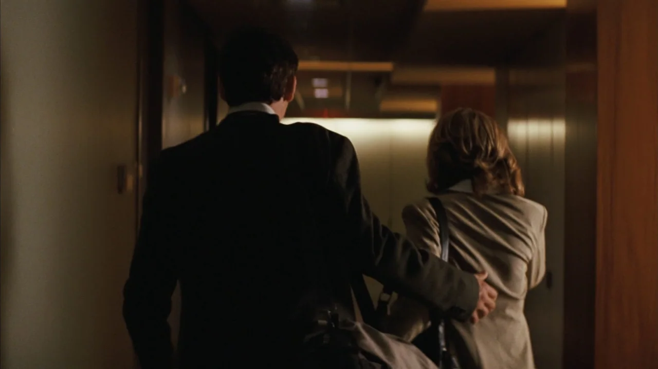 Duplicity (2009) movie still: medium — A dimly lit hotel hallway features dark wood paneling and beige-toned wall lights. A man in a dark suit leads…