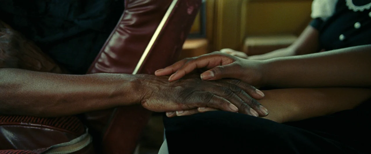 All Dirt Roads Taste of Salt (2023) movie still: closeup — An aged hand with prominent veins rests beneath a younger smooth hand. The hands are…