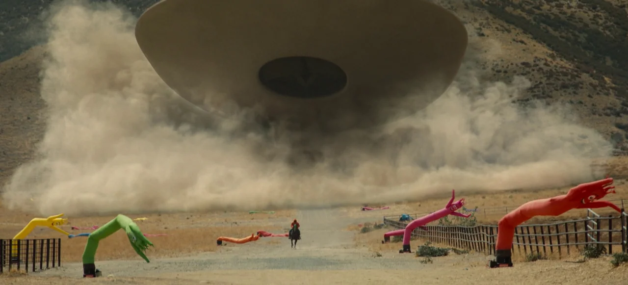 Nope (2022) movie still: wide — A dusty, sun-bleached landscape features a massive disc-shaped object hovering above a dirt road. A rider on…