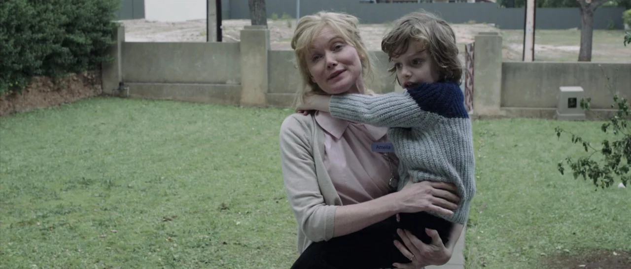 The Babadook (2014) movie still: medium — A woman stands in a grassy backyard near a low concrete wall. She holds a young boy in her arms while…