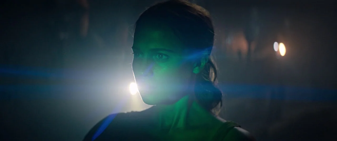 Tomb Raider (2018) movie still: closeup — A dark, cramped space with textured walls and faint distant light sources. A woman stands in the…