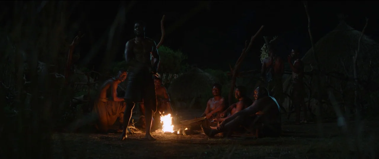The Woman King (2022) movie still: wide — A group of shirtless men gathers at night in a small, traditional outdoor settlement. They sit and stand…