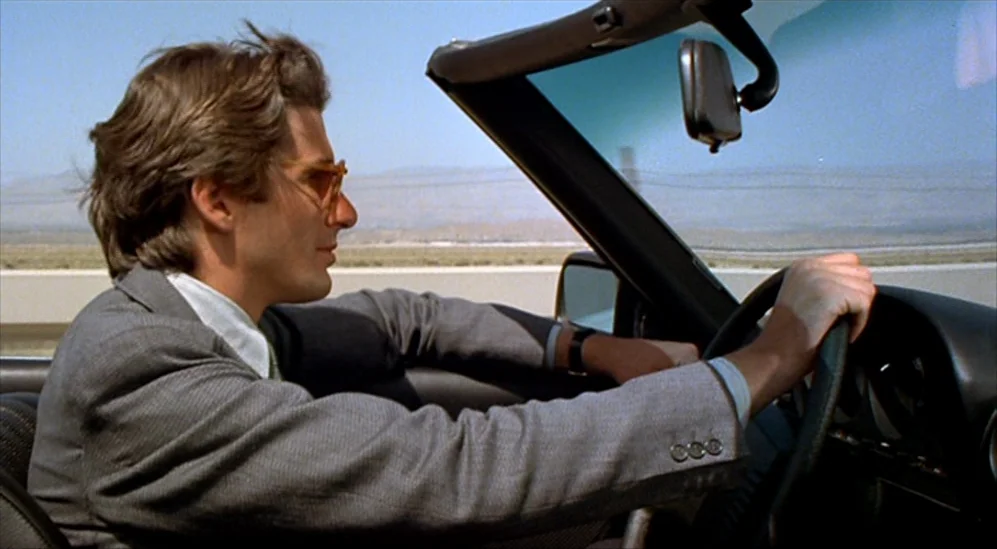 American Gigolo (1980) movie still: medium — A man drives a convertible along a flat desert highway under a clear sky. He wears a grey suit and…