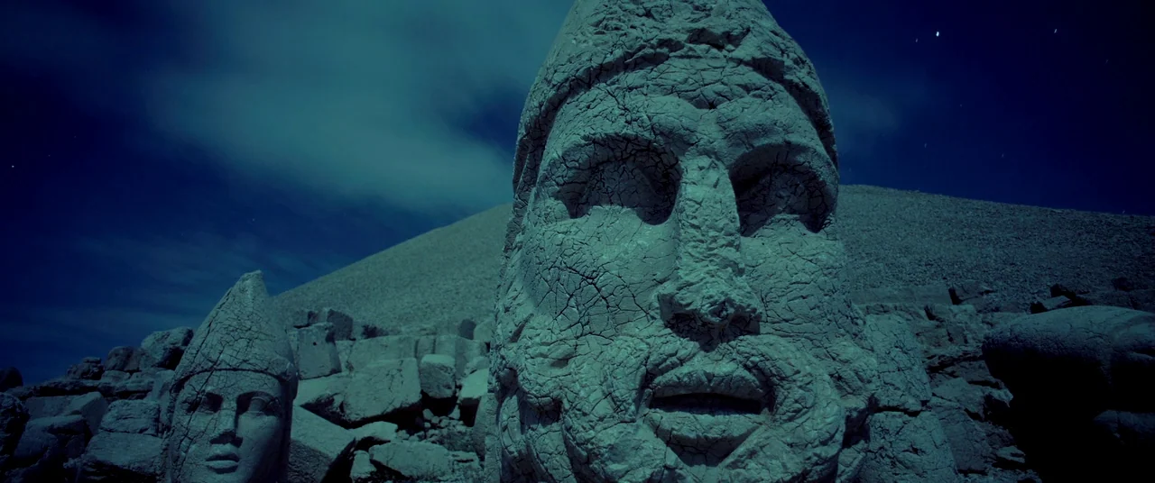 Samsara (2011) movie still: closeup — Massive stone heads of ancient statues sit on a rocky mountainside at night. A clear blue sky with stars…