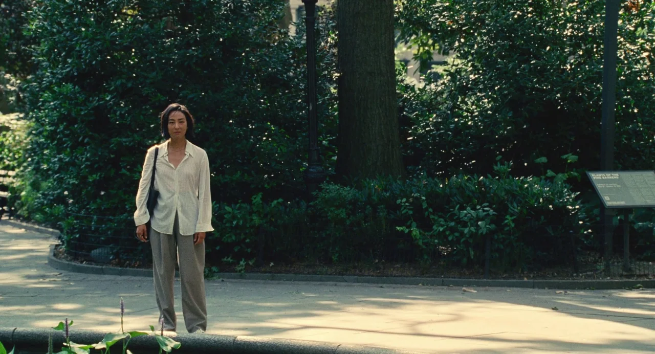 Past Lives (2023) movie still: fullbody — A paved path runs through a shaded urban park with dense green shrubbery behind. A woman wearing a…