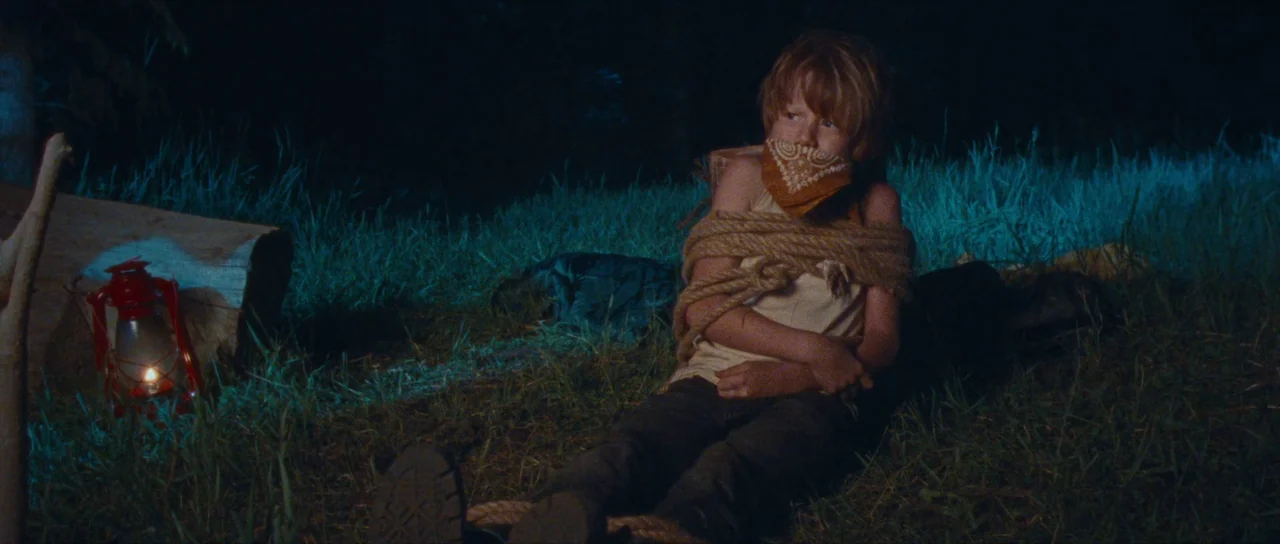 Riddle of Fire (2023) movie still: medium — A night campsite is illuminated by the dim glow of a red oil lantern. A young boy sits tied up on the…