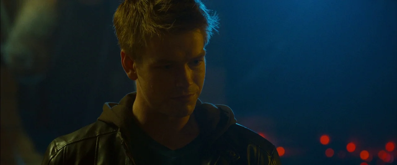 The Guest (2014) movie still: medium — A dark, blurred nightclub interior features a man standing in the foreground. He looks down with a neutral…