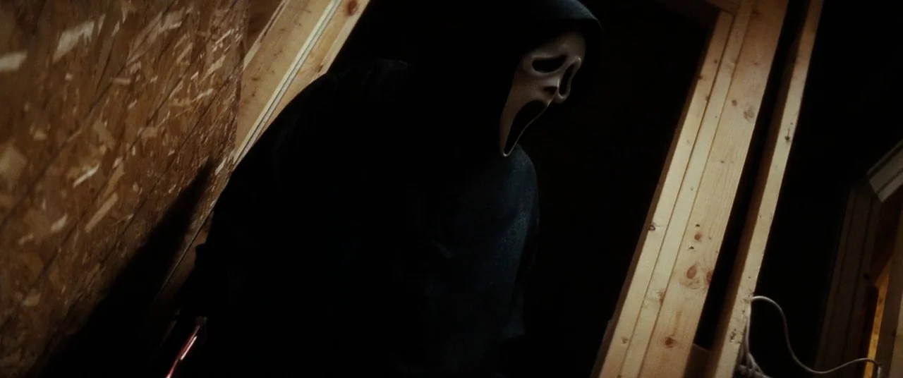 Scream 4 (2011) movie still: medium — An unfinished room with exposed wooden wall studs and plywood paneling in the background. A figure in a dark…